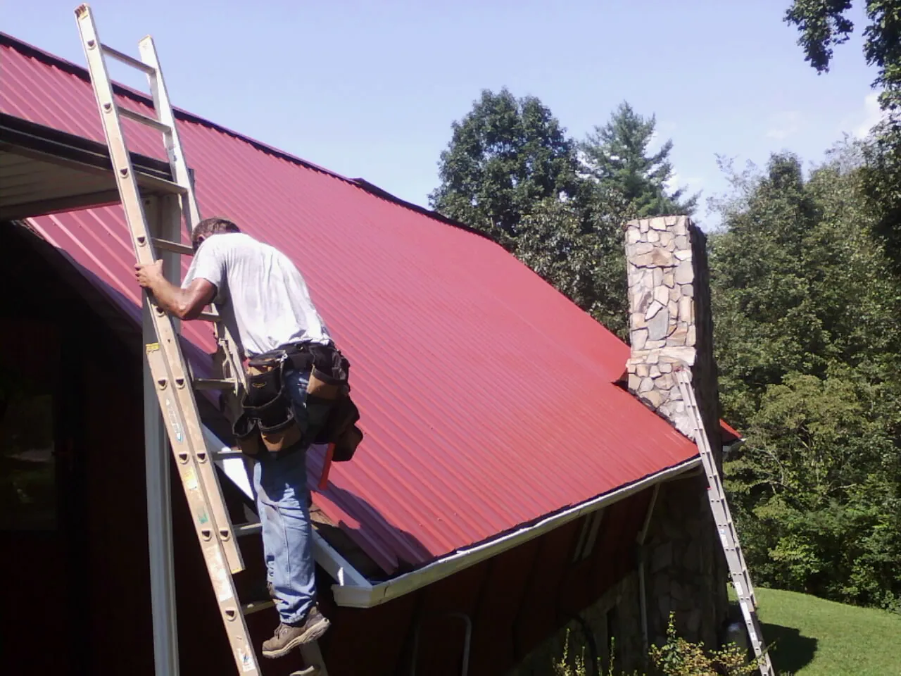 Roofer on a ladder beside a red metal roof and stone chimney — Soffit & Fascia Repair by Roofing Brothers in Buckholts